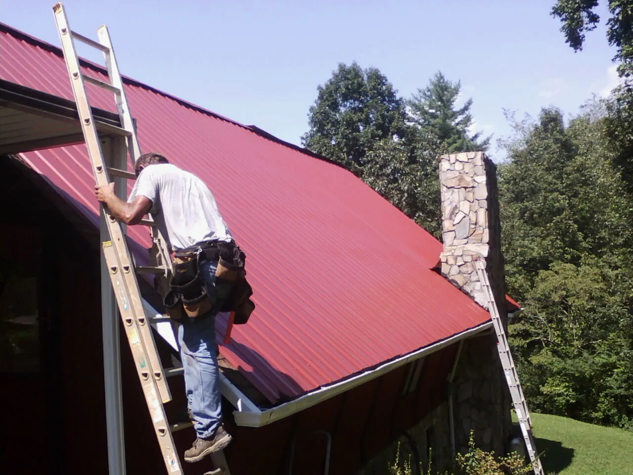 Roofer on a ladder beside a red metal roof and stone chimney — Soffit & Fascia Repair by Roofing Brothers in Graham
